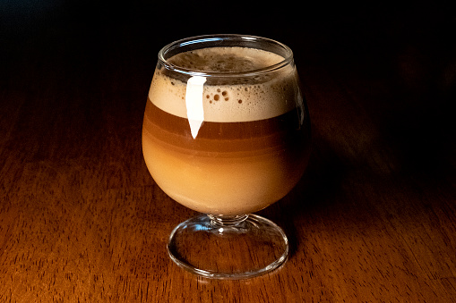 A cappuccino  in a small glass on a wood grain table with dark background.