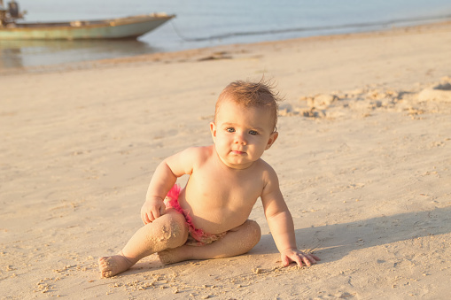 Cute little baby girl is sitting on a sandy beach near to sea in sunset sunlight.