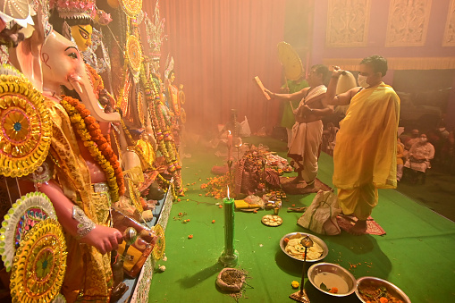 Howrah, India -October 13, 2021 : Hindu Priests worshipping Goddess Durga with ghanta, chamor and hand fan. Ashtami puja aarati - sacred Durga Puja ritual - shot at night.