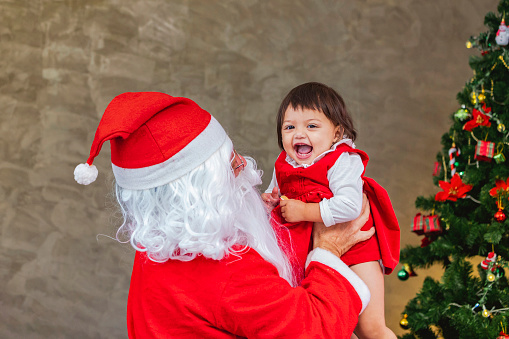 Santa Claus is lifting happy little toddler baby girl up and laughing cheerfully with fully decorated christmas tree on the back for season celebration