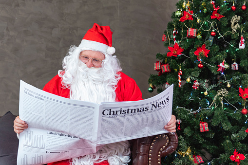 Santa Claus is reading Christmas news from the newspaper while sitting on the sofa couch by the christmas tree for long holiday economics situation and accident traffic transport report