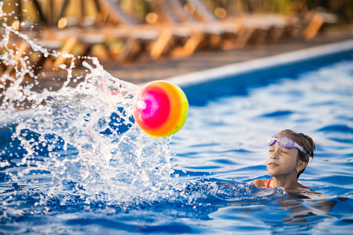 A young girl in a bright leopard swimsuit, swims with a bright colored inflatable ball in a deep blue pool with clear transparent water on a warm sunny summer evening