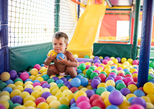 Little cheerful funny boy in a diaper with an inflatable green striped ball in his hands is playing in playroom in the weekend, enjoying warm bright summer days