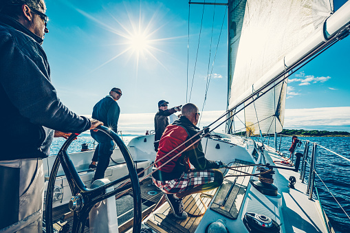 Mature man steering a private yacht while on vacation. Bearded yachtsman standing on his sailing boat looking at distance.