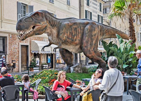 Albenga, Italy - May 14, 2022: A replica of a dinosaur in a piazza in Albenga, Italy. In the foreground people sitting at tables of an outdoor cafe.