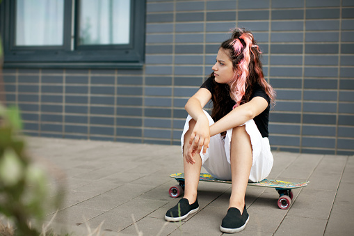 Teenager girl sits on skateboard. Hipster Teen model skateboarder with long pink hair on gray background
