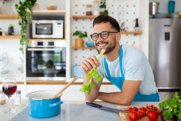 istock Handsome smiling young man leaning on kitchen counter with vegetables and looking away. Portrait of happy casual guy in apron leaning on steel counter in the kitchen with ingredients on it. 1412160928