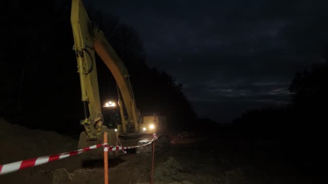 An excavator with headlights on stands at a construction site in the dark. Night shift work. Safety engineering, industry.