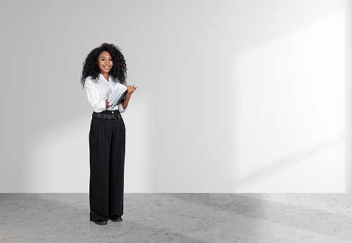 African businesswoman take note in business journal, looking at the camera and smiling, full length on grey concrete floor. Copy space empty white wall.