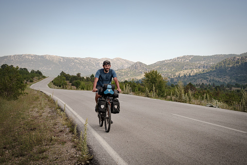 Photographed from the front view. Model with bicycle helmet drives on the curved asphalt road. Adherent to the adventurous spirit, high vitality, Mountains in the background.He is 30 years old, doing a camping bike tour. There are various bags on his bike, around 60 liters in total.Mediterranean region Antalya.