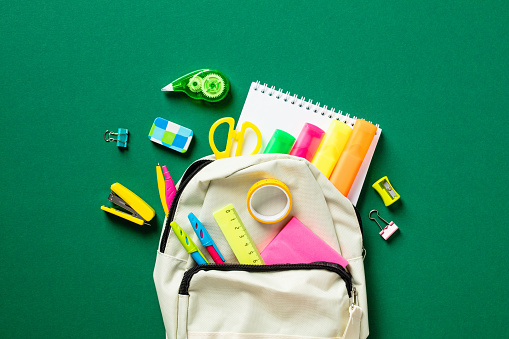 Backpack with colorful school supplies on green background. Back to school concept. Flat lay, top view, copy space.