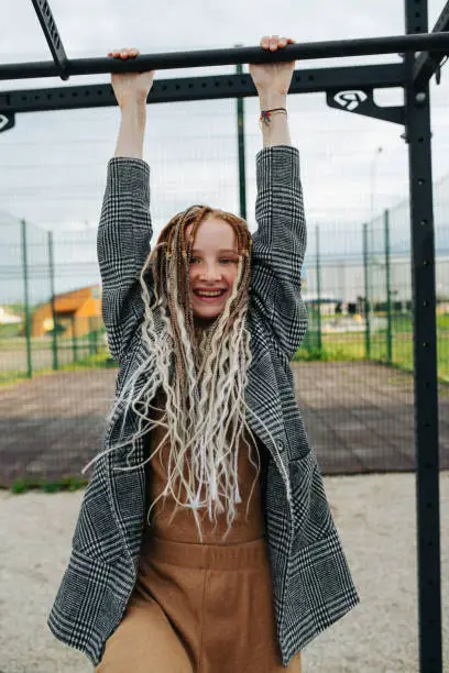 Freckled teenage girl with dreads hanging on a high bar on the sportsground. Looking at the camera.