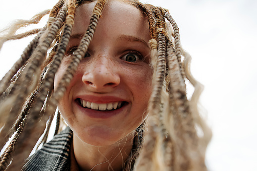 Creepy happy close up face of a freckled teenage girl with dreads. Upward angle. Against a grey sky.