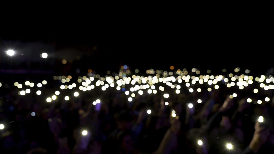 Defocused spectators in hall with lights waving in the dark. Action. Audience holding phones with shining flashlights during the concert, romantic atmosphere.