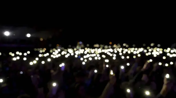 Defocused spectators in hall with lights waving in the dark. Audience holding phones with shining flashlights during the concert, romantic atmosphere.