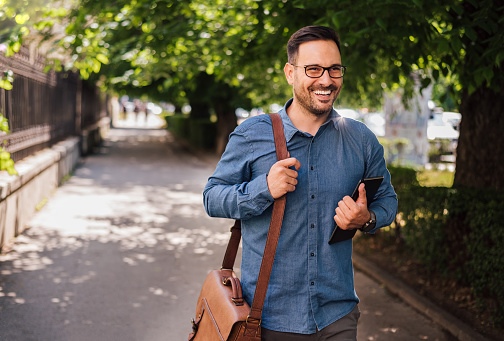 Smiling young male professional with digital tablet and bag looking away while walking on sidewalk.