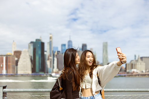 Two Asian tourist girls, students walking Brooklyn Bridge park with a view of lower Manhattan. They are taking selfie for a memory. Dumbo, Brooklyn, New York
