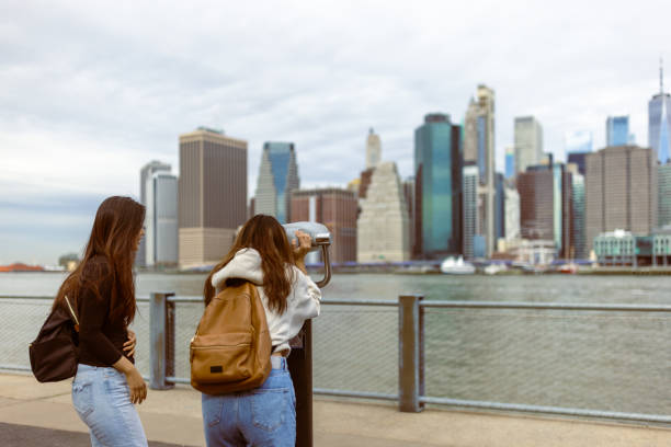 Two Asian tourist girls, students walking Brooklyn Bridge Park. They are observing  Lower Manhattan using public binocular. Dumbo, Brooklyn, New York