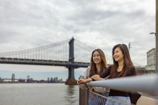 Two Asian tourist girls, students walking Brooklyn Bridge park with a view of lower Manhattan. Dumbo, Brooklyn, New York