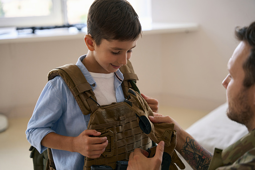 Man of arms demonstrating gloves to his son, who wearing military tactical vest