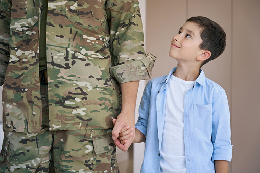 Proud boy looking up at his soldier father while holding his hand at home