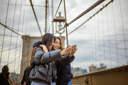 Two Asian tourist girls, students walking Brooklyn Bridge. They are taking selfie for a memory. Dumbo, Brooklyn, New York