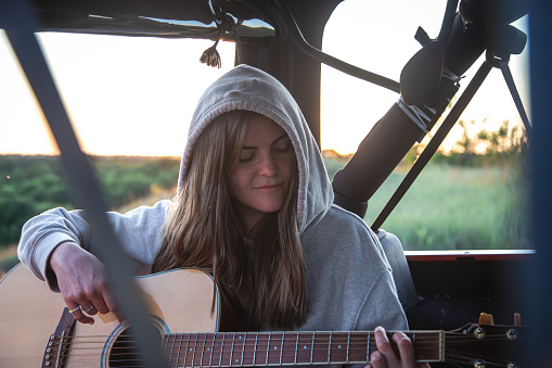 A young woman playing acoustic guitar in car trunk in nature, road trip concept.