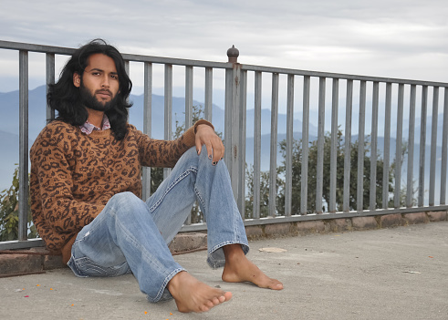 A good looking Indian young man with long hair and beard looking at camera while sitting with leaning on safety barrier against the background of mountains
