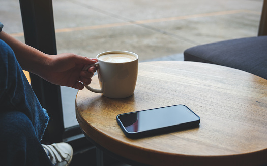 Closeup image of a woman holding and drink hot coffee with mobile phone on the table in cafe