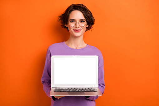 Photo of charming lady hold demonstrate empty space netbook display isolated on orange color background.