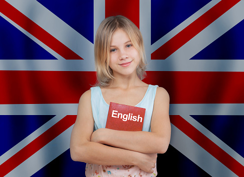 Smiling child girl holding english book against UK flag background. Learn English language concept.
