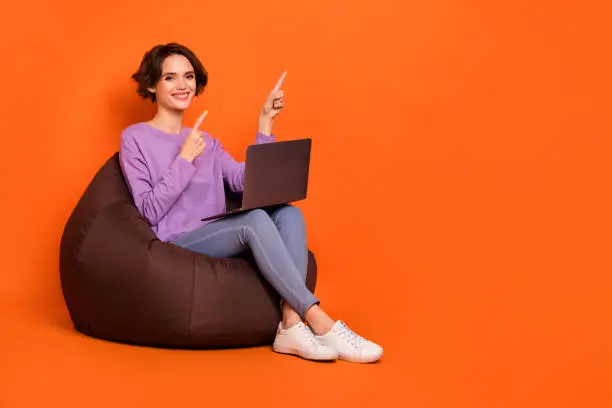 Full length photo of cute shiny lady wear violet sweatshirt sitting bean bag typing device pointing empty space isolated orange color background.