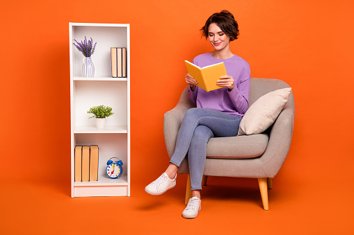 Full length photo of shiny funny girl dressed purple pullover reading story sitting armchair isolated orange color background.