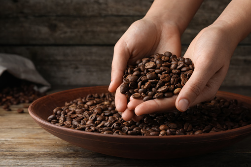 Woman taking pile of roasted coffee beans from bowl at wooden table, closeup