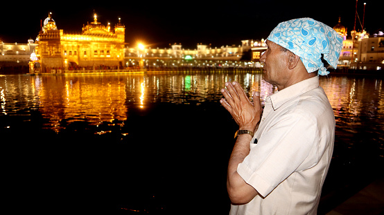 Senior man of Indian ethnicity standing in Golden temple at night and he praying to god. Shoot location Golden Temple, Amritsar, Punjab, India.