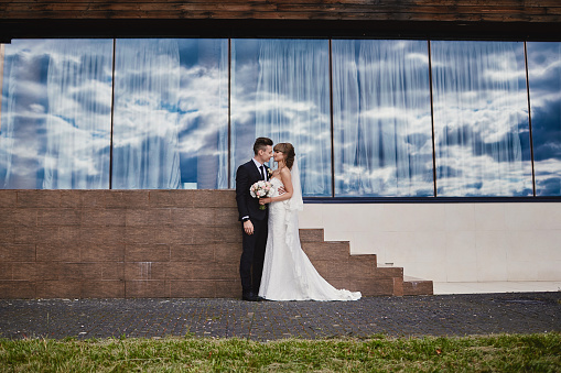 Young couple in love bride and groom posing outdoors near the large panoramic window. woman in white long dress and bouquet with flowers hugging man in black suit. wedding day.