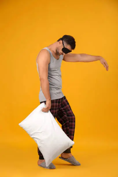 Man with pillow and eye mask in sleepwalking state on yellow background