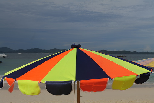 A colorful sunshade umbrella at a popular tourist destination island of Thailand (Khai). In the background you see the azure water and white sand of an idyllic beach. The sky is cloudy, a monsoon rain is approaching.