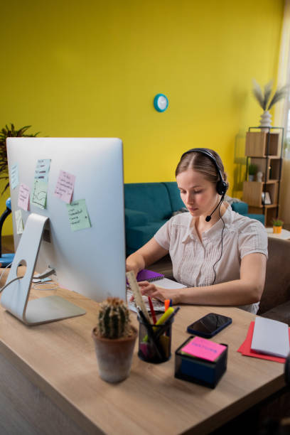 Portrait view of focused young woman in headset with microphone sitting on sofa, involved in video call conversation. Female having online meeting workshop with colleagues Portrait view of focused young woman in headset with microphone sitting on sofa, involved in video call conversation. Female having online meeting workshop with colleagues. school receptionist stock pictures, royalty-free photos & images