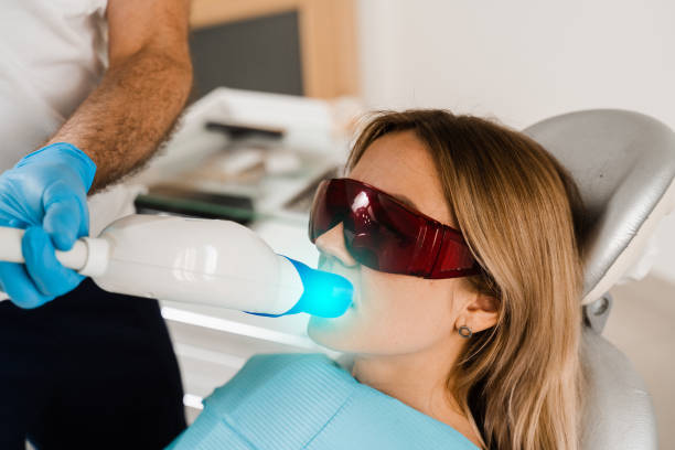 istock Artificial teeth whitening. Dental ultraviolet whitening treatment with light, fluoride and laser. Woman in red protective glasses patient do UV teeth whitening procedure. 1412232049