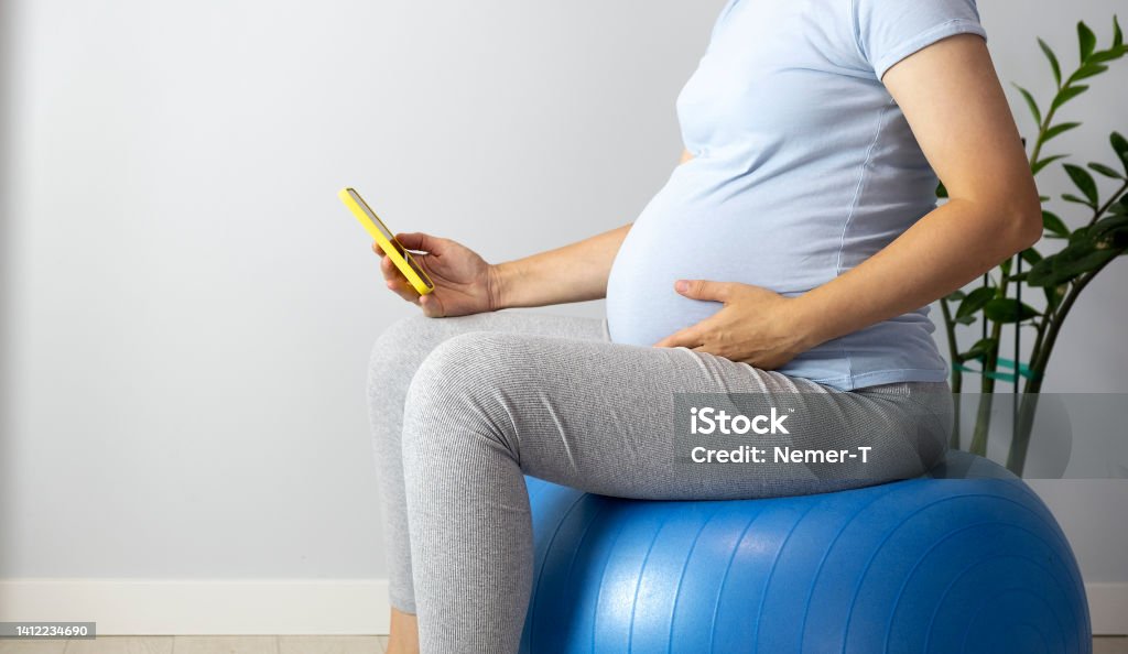 Pregnant woman on fitball Pregnant woman feel contractions sitting on fitball and use mobile time counter to count contraction. Smartphone application for pregnancy and childbirth. Fitness Ball Stock Photo