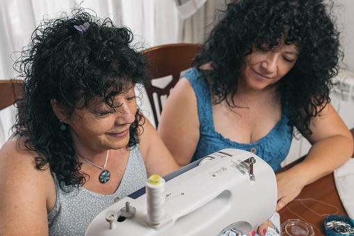 Mother and daughter doing sewing and tailoring work at home. Older lady teaching her adult daughter how to use the automatic sewing machine. Diagonal view.