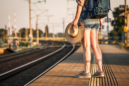 Traveler with backpack and hat waiting for train at railroad station platform. Woman tourist going to summer vacation alone