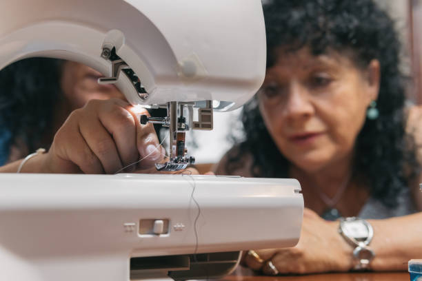 istock Older woman watching how her daughter performs the sewing and tailoring work she has just taught her. Curly-haired lady supervising her daughter threading the thread on the sewing machine threader. 1412238302