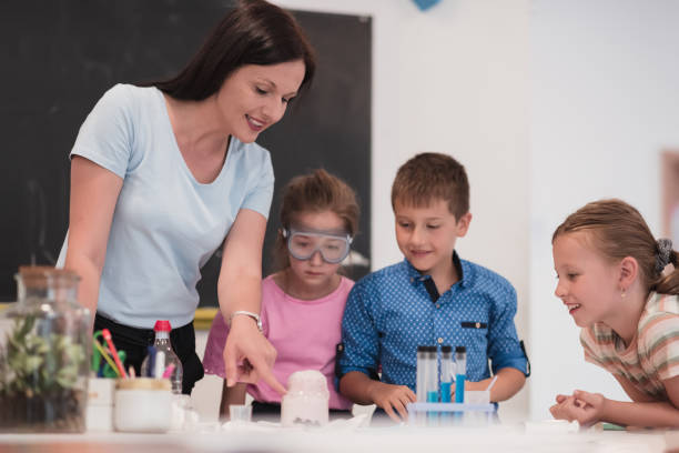 istock Elementary School Science Classroom: Enthusiastic Teacher Explains Chemistry to Diverse Group of Children, Little Boy Mixes Chemicals in Beakers. Children Learn with Interest 1412243031
