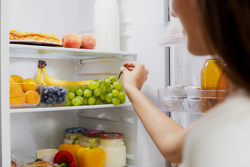 Woman hand taking, grabbing or picks up green bunch of grapes out of open refrigerator shelf or fridge drawer full of fruits, vegetables, banana, peaches, yogurt. Healthy food diet, lifestyle concept