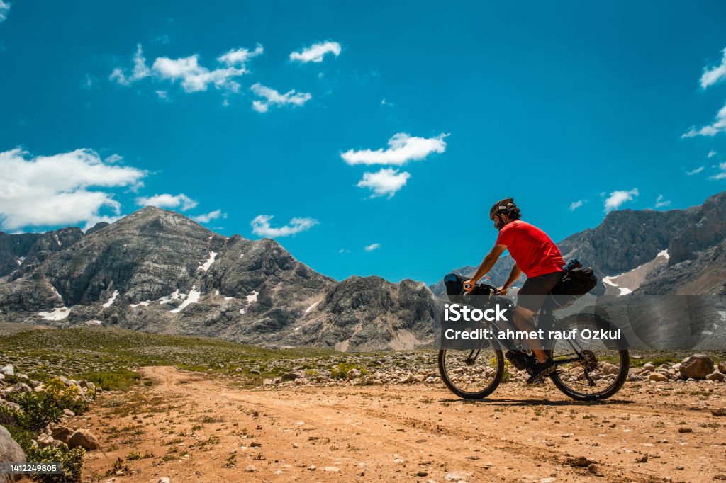 Triangle less snowy imposing mountain, dirt road bike tour He is 30 years old, doing a camping bike tour. There are various bags on his bike, around 60 liters in total.Mountainous region less cloudy weather nature view. Tour cyclist walking on dirt road. Mountain triangle is snowy in places. The spirit of adventure is high. Mediterranean region Taurus Mountains Antalya. Cycling Stock Photo