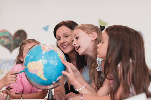 Female teacher with kids in geography class looking at a globe. Side view of a group of diverse happy school kids with a globe in the classroom at school. High quality photo