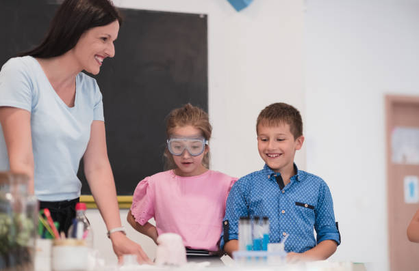 istock Elementary School Science Classroom: Enthusiastic Teacher Explains Chemistry to Diverse Group of Children, Little Boy Mixes Chemicals in Beakers. Children Learn with Interest 1412267280