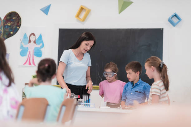 istock Elementary School Science Classroom: Enthusiastic Teacher Explains Chemistry to Diverse Group of Children, Little Boy Mixes Chemicals in Beakers. Children Learn with Interest 1412268246